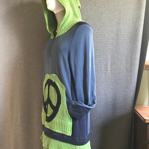 Könnte beinhalten: Ein blauer und grüner Kapuzenpullover mit einem Peace-Zeichen auf der Vorderseite. Der Pullover hat einen Reißverschluss und einen gekräuselten Saum.