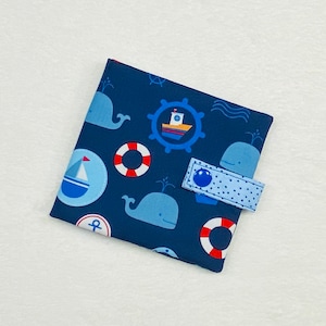 Könnte beinhalten: Ein marineblaues Stoffstück mit maritimem Thema. Der Stoff zeigt hellblaue Wale, Segelboote, Rettungsringe und Steuerräder. Ein hellblauer Riemen mit einem blauen Knopf sichert den Artikel. Der Hintergrund ist weiß.