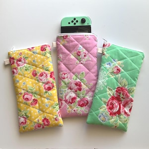 May include: Three quilted fabric cases with floral patterns. One is yellow with white polka dots, another is pink, and the third is green. A mint green game console is partially visible inside the pink case.