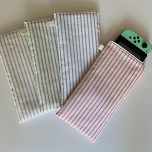 May include: Four fabric cases with vertical striped patterns in various colors. One case is open, holding a mint green handheld gaming device. The cases have a rectangular shape with a zipper closure and a small fabric loop.