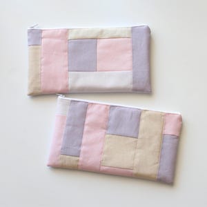 May include: Two rectangular, zippered pouches with a patchwork design. The pouches are made of fabric in shades of pink, lavender, and beige. The zippers are white. The pouches are arranged on a white surface.