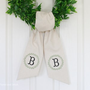 Pu&ograve; includere: Un fiocco per ghirlanda in tessuto bianco con una ghirlanda verde e la lettera "B" ricamata su ogni lato.