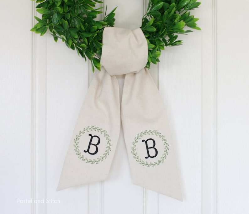 Pu&ograve; includere: Un fiocco per ghirlanda in tessuto bianco con una ghirlanda verde e la lettera "B" ricamata su ogni lato.