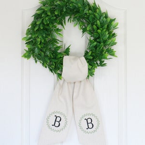 Pu&ograve; includere: Una ghirlanda verde con un fiocco di stoffa bianco legato intorno. Il fiocco ha la lettera "B" ricamata su ogni lato all'interno di una ghirlanda di foglie.