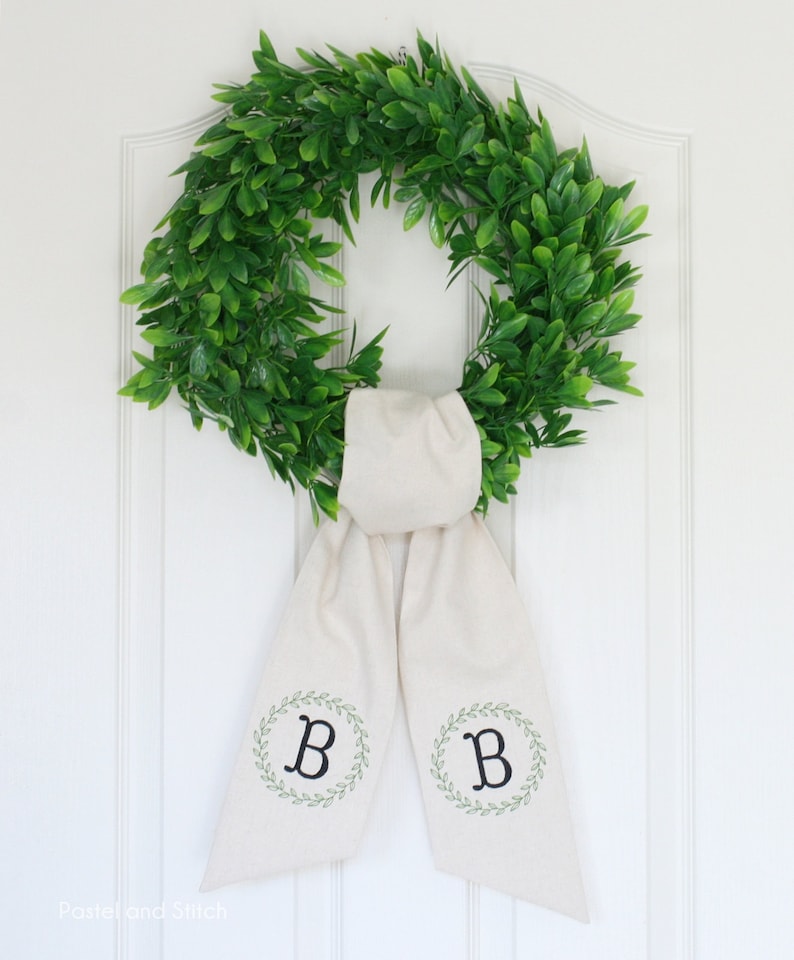 Pu&ograve; includere: Una ghirlanda verde con un fiocco di stoffa bianco legato intorno. Il fiocco ha la lettera "B" ricamata su ogni lato all'interno di una ghirlanda di foglie.
