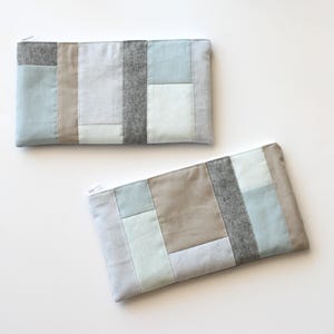 May include: Two rectangular zippered pouches with a patchwork design. The pouches feature a mix of light blue, gray, and beige fabric panels. The top pouch is oriented horizontally, while the bottom pouch is at an angle.