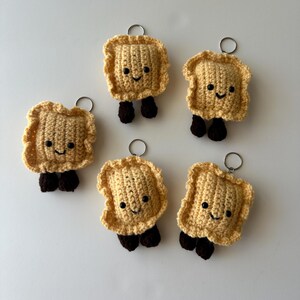 May include: Five handmade crochet ravioli keychains. Each keychain features a smiling face, tan body, ruffled edges, and brown feet. The keychains are attached to silver-colored key rings.