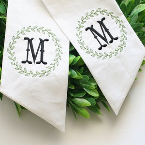 Pu&ograve; includere: Due stendardi di tessuto bianco con ghirlande ricamate in verde e una lettera "M" ricamata in nero al centro di ogni ghirlanda. Gli stendardi sono esposti su uno sfondo di fogliame verde.