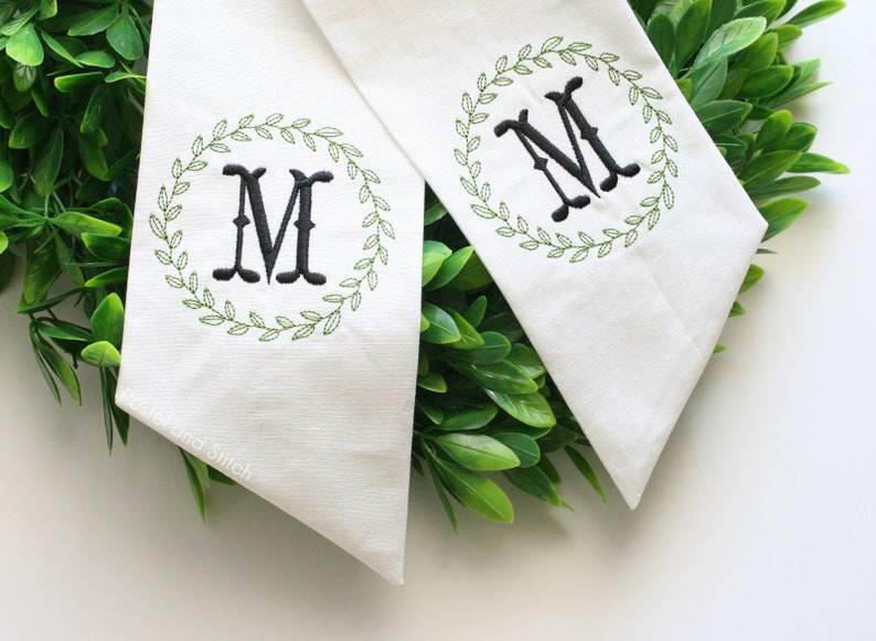Pu&ograve; includere: Due stendardi di tessuto bianco con ghirlande ricamate in verde e una lettera "M" ricamata in nero al centro di ogni ghirlanda. Gli stendardi sono esposti su uno sfondo di fogliame verde.