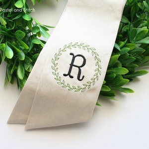 Pu&ograve; includere: Uno stendardo in tessuto beige con una ghirlanda verde ricamata attorno alla lettera "R" in nero. Lo stendardo &egrave; parzialmente avvolto attorno a una ghirlanda verde.