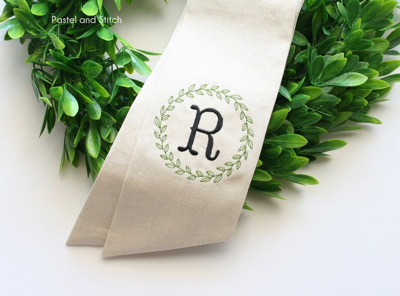 Pu&ograve; includere: Uno stendardo in tessuto beige con una ghirlanda verde ricamata attorno alla lettera "R" in nero. Lo stendardo &egrave; parzialmente avvolto attorno a una ghirlanda verde.