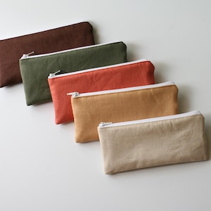 May include: Five rectangular pencil cases in shades of brown, green, orange, tan, and beige. Each case has a white zipper and a white trim. The cases are arranged diagonally on a white surface.