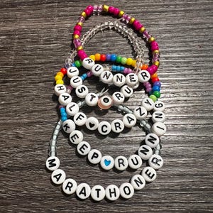 May include: A set of colorful beaded bracelets with the words "Runner", "Just Run", "Half Crazy", "Keep Running", and "Marathoner" spelled out on them. The bracelets are made with small, round beads in various colors, including pink, purple, blue, green, yellow, and white. The words are spelled out in black letters on white beads.