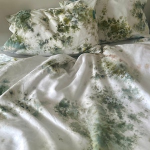 May include: A white duvet cover and two pillowcases with a green and blue tie-dye pattern. The fabric has a soft, wrinkled texture.