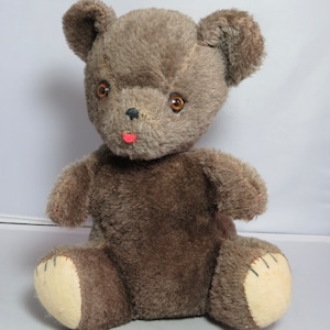 May include: A brown teddy bear with a pink nose and brown button eyes. The bear is sitting with its paws in front of it.
