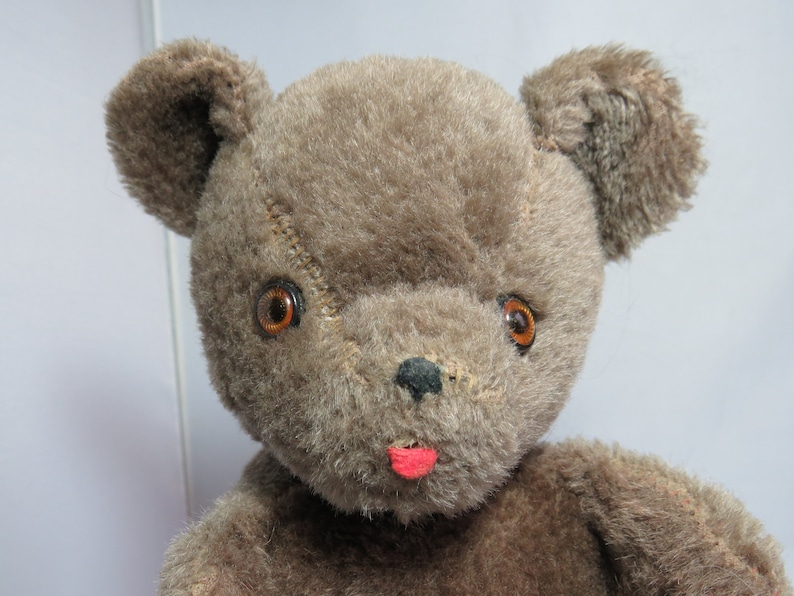 May include: A brown teddy bear with large, round, brown eyes and a pink tongue sticking out. The bear has a stitched seam across its face and a worn, fuzzy texture.