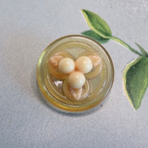 May include: A yellow button with a raised design of three white pearls and leaves. The button has a concentric circle pattern.