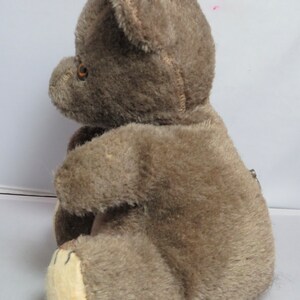 May include: A brown plush teddy bear with a beige belly and brown eyes. The bear is sitting on a white surface.