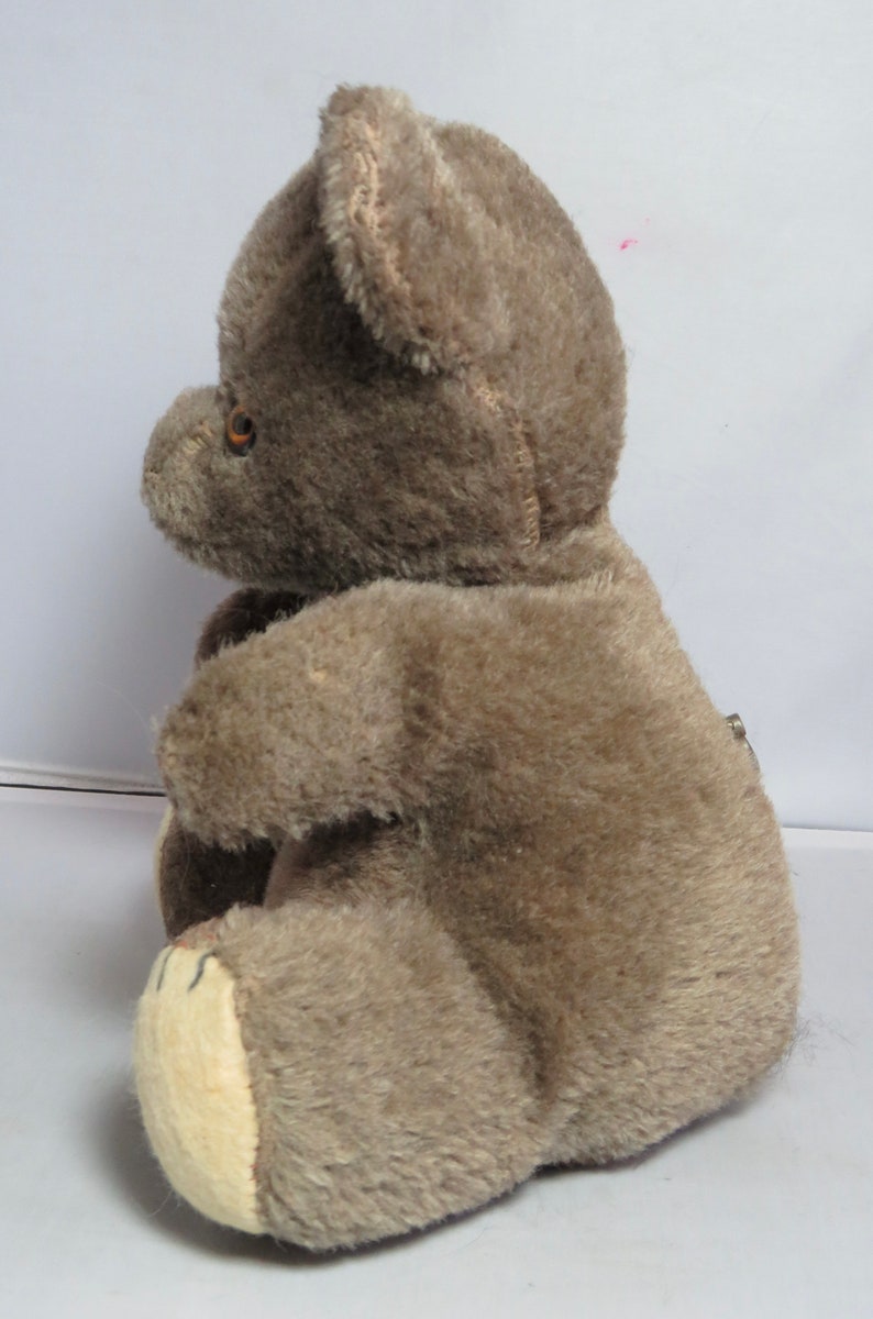 May include: A brown plush teddy bear with a beige belly and brown eyes. The bear is sitting on a white surface.