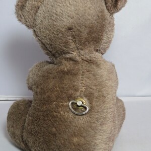 May include: A brown plush teddy bear with a wind-up key on its back. The bear is sitting on a white surface.