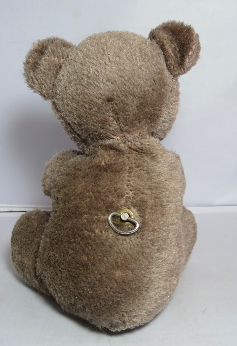 May include: A brown plush teddy bear with a wind-up key on its back. The bear is sitting on a white surface.