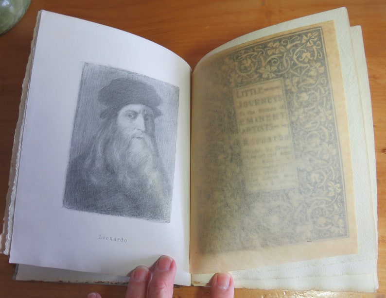 May include: An open book with a black and white illustration of Leonardo da Vinci on the left page. The right page has a decorative border with the text "Little Journeys to the Homes of Great Artists" in the center.