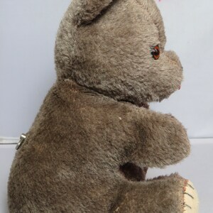 May include: A brown plush teddy bear with a keywind mechanism in the back. The bear is sitting with its right leg extended and its left leg tucked under its body. The bear has brown fur and a stitched nose.