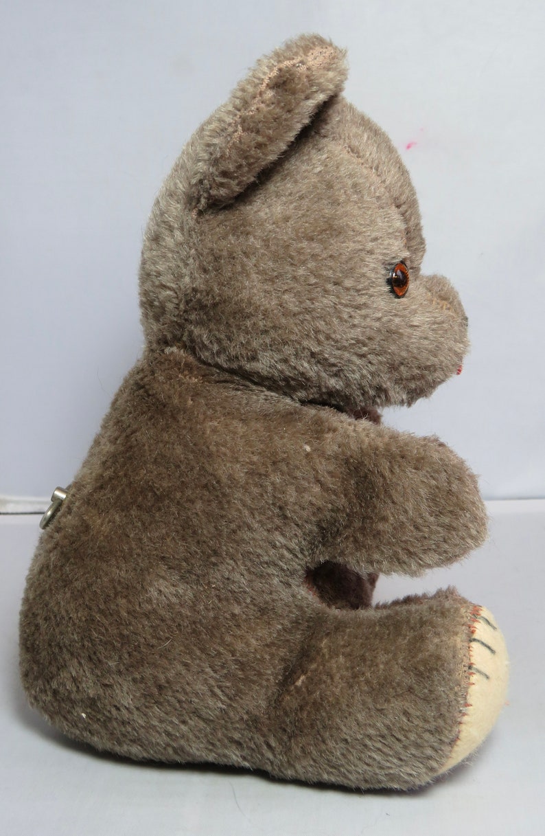 May include: A brown plush teddy bear with a keywind mechanism in the back. The bear is sitting with its right leg extended and its left leg tucked under its body. The bear has brown fur and a stitched nose.