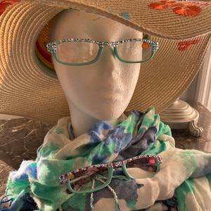 Può includere: Una sciarpa blu turchese con un motivo floreale bianco e blu. La sciarpa è drappeggiata su una testa di manichino bianca che indossa un cappello di paglia a tesa larga con un bordo arcobaleno e occhiali in strass blu turchese.