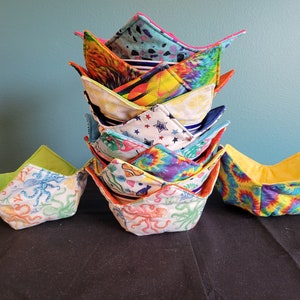 May include: A stack of colorful fabric bowl cozies with various patterns, including tie-dye, stars, and sea creatures. The cozies are folded in a triangular shape and are made of soft, quilted fabric.