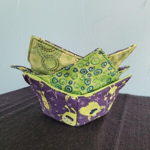 May include: Three fabric bowl cozies. The top cozie is green with a swirling pattern. The middle cozie is green with a circular pattern. The bottom cozie is purple with a green alien pattern.