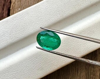 Puede incluir: Una esmeralda ovalada de color verde vibrante sostenida por unas pinzas. La esmeralda tiene un corte facetado y está colocada sobre un fondo blanco.