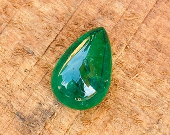 Puede incluir: Un cabujón de piedra preciosa verde en forma de lágrima, con una superficie lisa y pulida. La piedra está colocada sobre un fondo de madera.