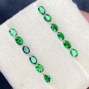 May include: A collection of vibrant green gemstones, including oval and pear-shaped cuts, arranged on a white fabric. The gemstones exhibit a rich, emerald-like color and high clarity, suggesting quality craftsmanship. Ideal for jewelry making or gemstone collecting.