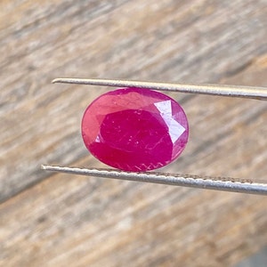 Può includere: Una gemma di un rosso vibrante e profondo, con taglio rotondo e sfaccettato. La gemma è tenuta da una pinzetta su uno sfondo di legno.