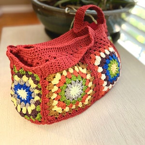 May include: A handmade red crochet bag with a handle. The bag features colorful granny square panels in shades of blue, green, yellow, and brown. The bag is sitting on a white surface.
