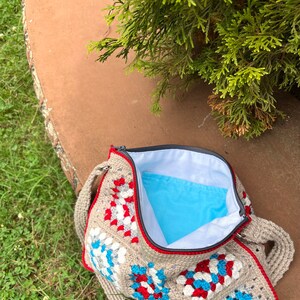 May include: A crocheted granny square bag with a red, white, and blue color scheme. The bag has a zipper closure and a long strap.