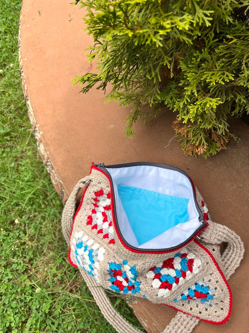 May include: A crocheted granny square bag with a red, white, and blue color scheme. The bag has a zipper closure and a long strap.