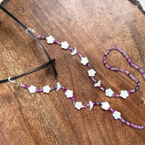 May include: A purple and white beaded glasses chain with flower and bird charms. The chain is attached to a pair of glasses.
