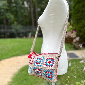 May include: A crocheted crossbody bag with a beige background and red, white, and blue squares. The bag has a zipper closure and a long strap.