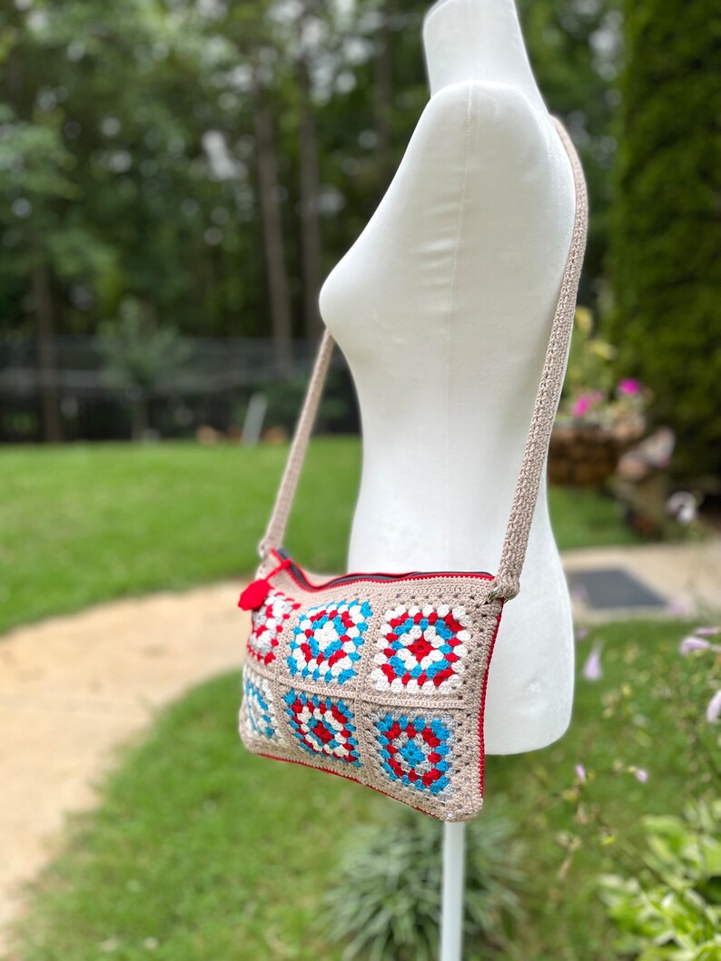 May include: A crocheted crossbody bag with a beige background and red, white, and blue squares. The bag has a zipper closure and a long strap.