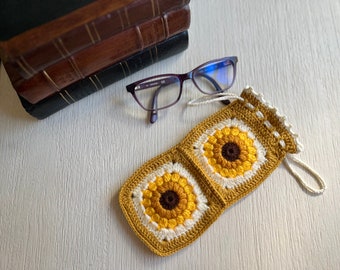 May include: A crocheted glasses case with a sunflower design in shades of yellow, white, and brown. The case has a drawstring closure and is made with a textured yarn.