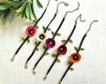 Può includere: Cinque collane in crochet nero con ciondoli a forma di fiore colorati. Ogni collana presenta un fiore di colore diverso con un centro giallo. I fiori sono realizzati in filo di crochet e hanno un aspetto delicato e femminile.