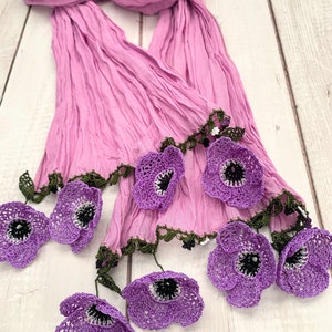 Pu&ograve; includere: Sciarpa lavanda con texture plissettata. Presenta fiori viola all'uncinetto con centri bianchi e neri e rifiniture verdi. La sciarpa &egrave; esposta su una superficie di legno chiaro.