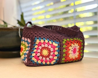 May include: A brown crocheted tote bag with colorful granny square designs. The squares feature pink, red, orange, yellow, green, blue, and brown yarn.