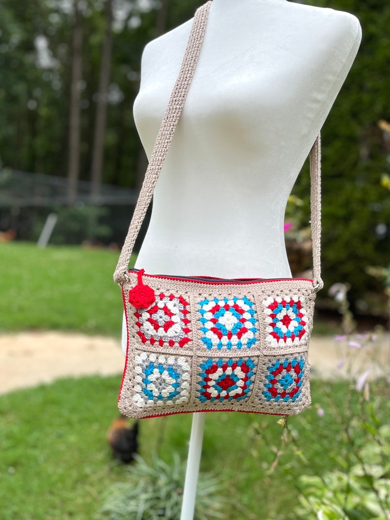 May include: A crocheted crossbody bag with a red, white, and blue granny square pattern. The bag has a beige strap and a red zipper.