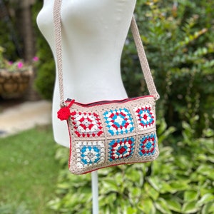 May include: A crocheted crossbody bag with a red, white, and blue geometric pattern. The bag has a long strap and a zipper closure.