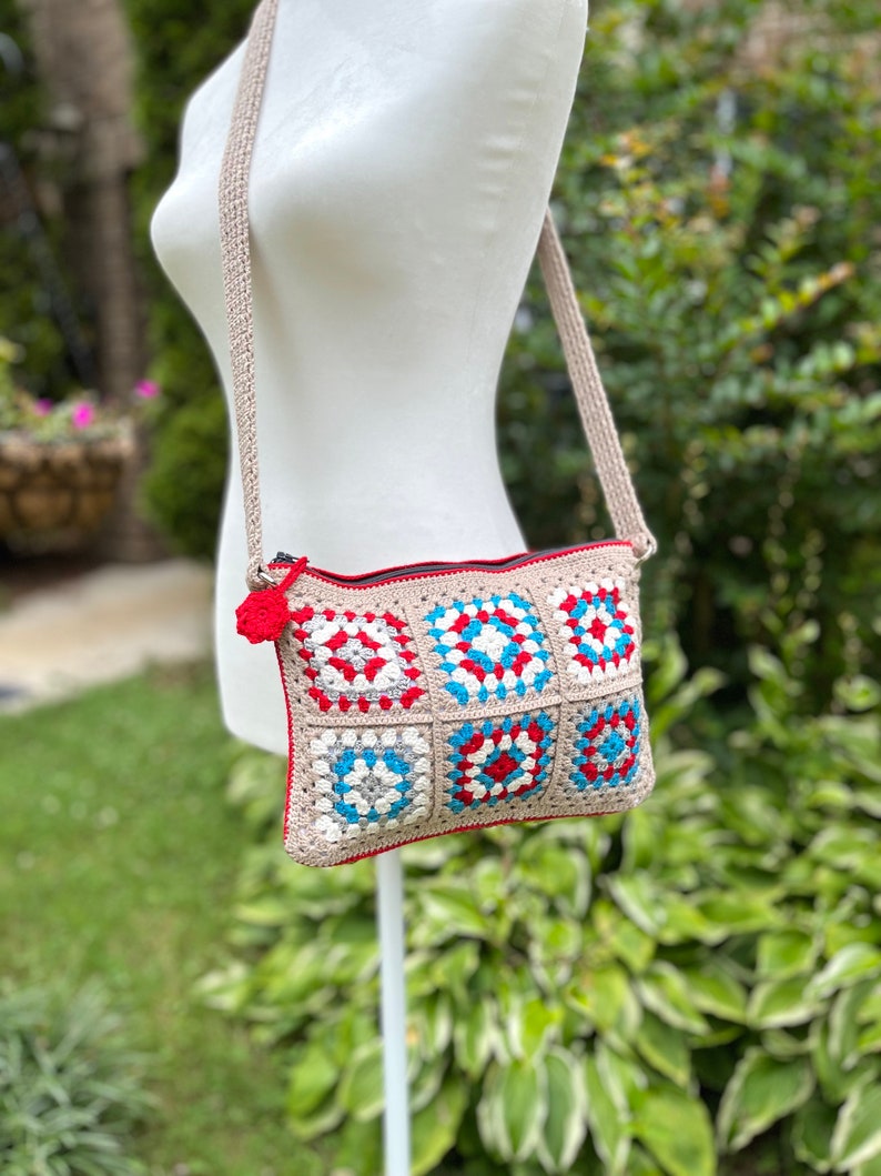 May include: A crocheted crossbody bag with a red, white, and blue geometric pattern. The bag has a long strap and a zipper closure.