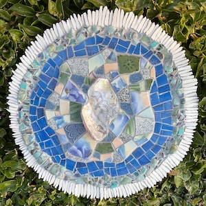 May include: A round mosaic art piece with a central oyster shell. The mosaic features shades of blue, green, and white tiles, with a white, textured border. The artwork is set against a backdrop of green foliage.