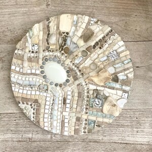 May include: A round mosaic art piece with a central oval shape. The mosaic is composed of various neutral-toned tiles, stones, and glass pieces in shades of white, beige, and gray. The design has a radial pattern.